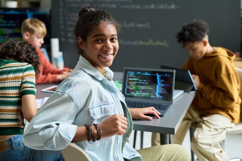 Teenage girl smiling at a desk with an open laptop in a classroom