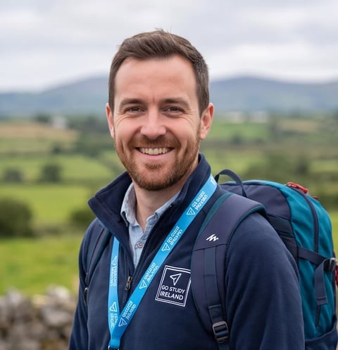 Man smiling at camera wearing navy jacket with lanyard and backpack in front of green rolling hills and mountains