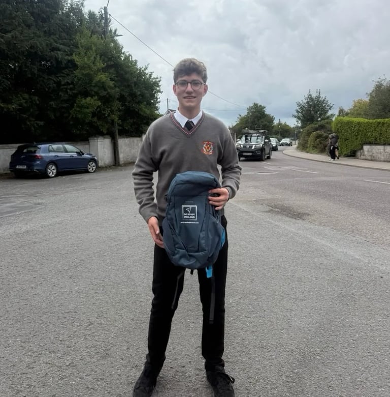 Young student wearing school uniform holding a blue backpack, standing on a street with parked cars in background
