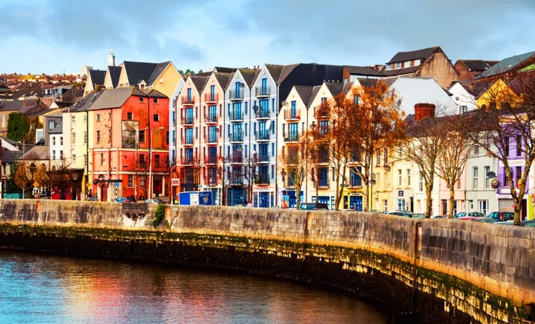 Colorful historic buildings along a waterfront with stone quay, featuring red, blue, and yellow Victorian architecture on a waterfront promenade