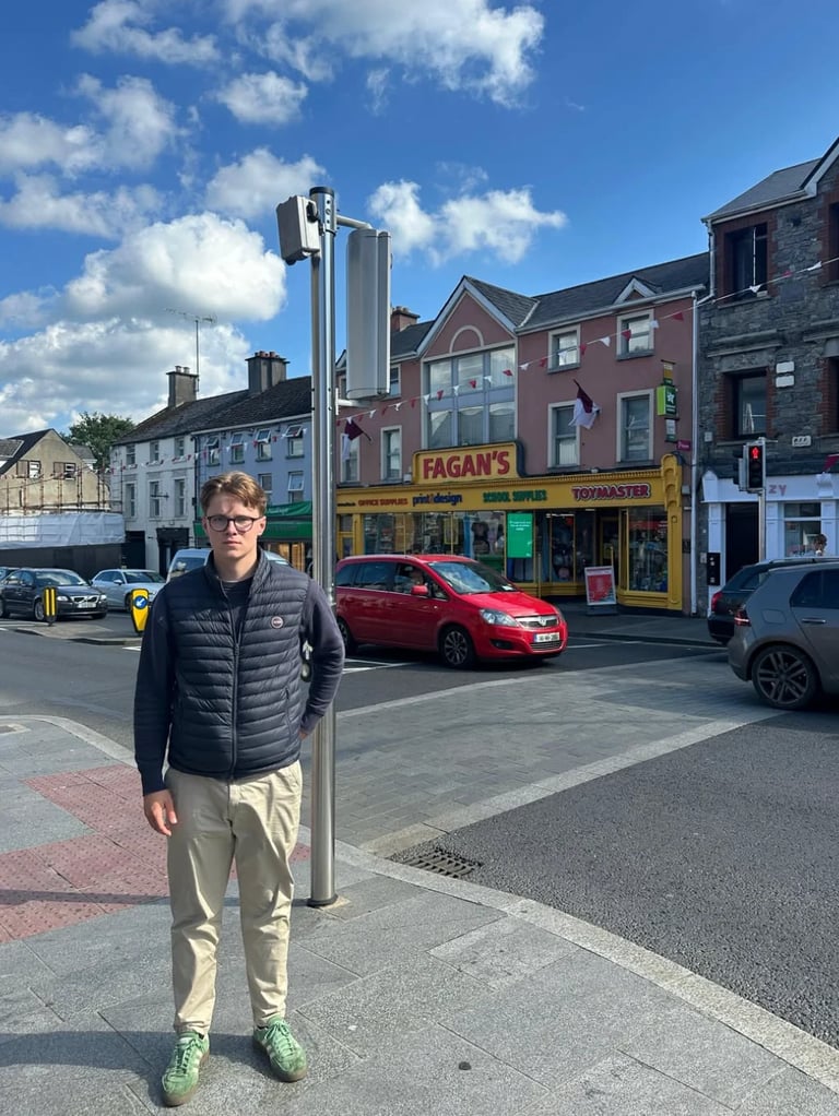 Young man standing on a town street next to a metal pole, with Fagans shop and parked cars visible in the background