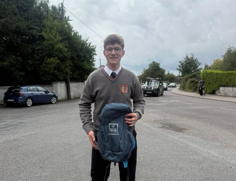 Young man in school uniform holding a blue backpack while standing in a parking lot with trees and vehicles in the background