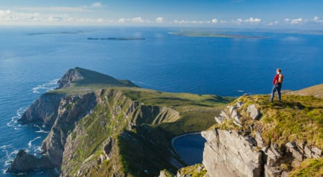Hiker standing on cliff edge overlooking green volcanic island with steep coastal cliffs and ocean views