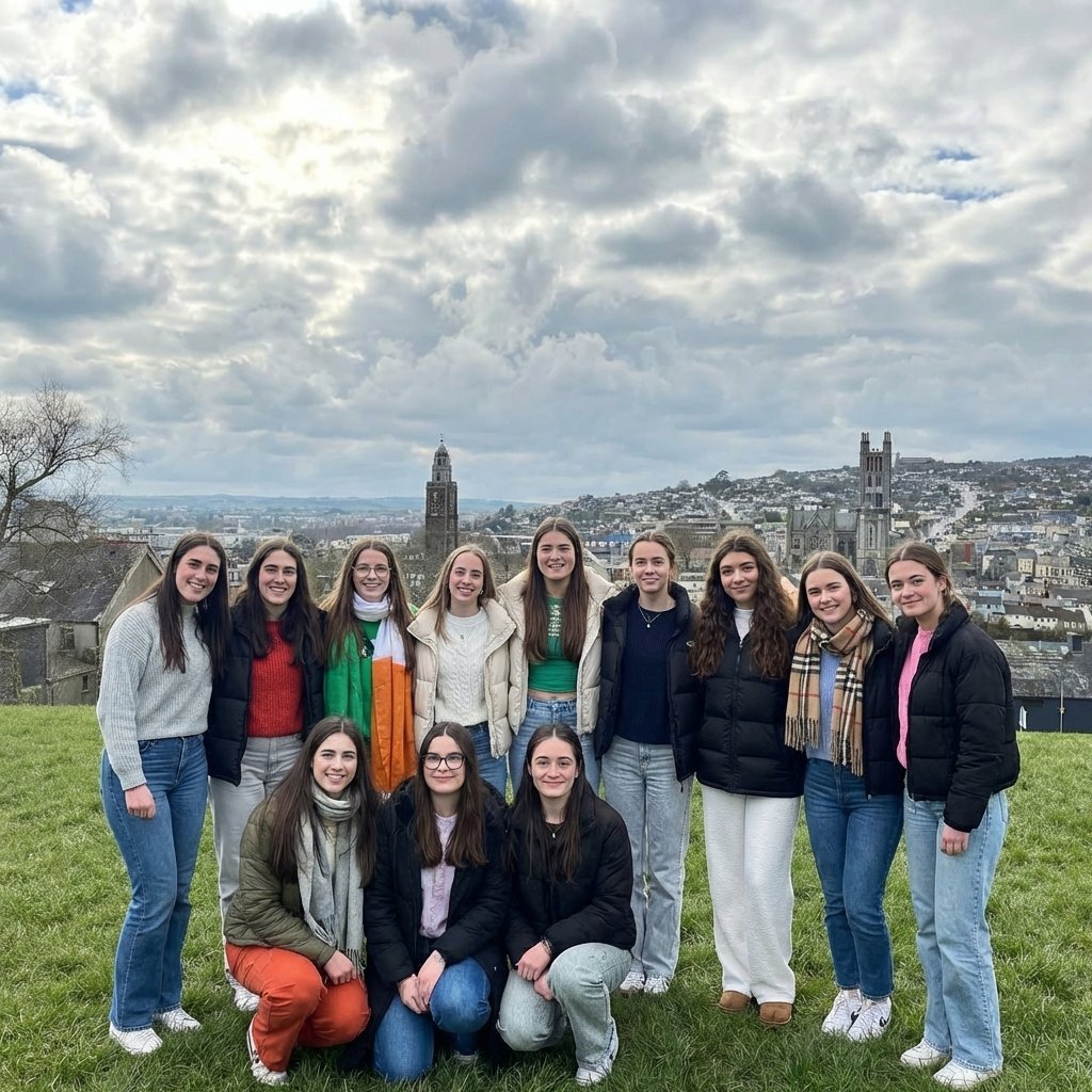 Group of 13 young women posing together on a grassy overlook with a historic European city skyline in the background