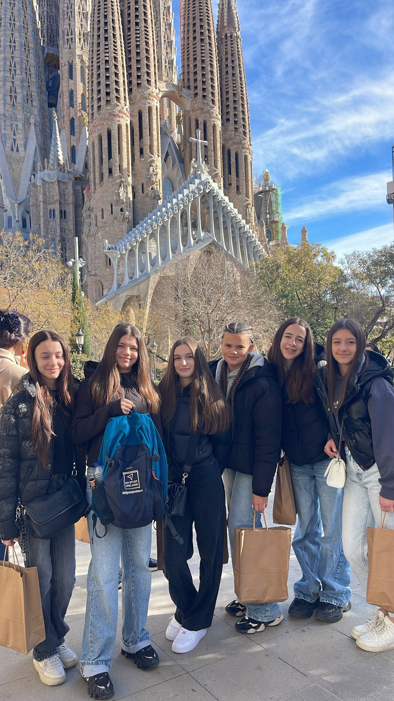 Six young women posing together in front of Sagrada Familia basilica in Barcelona, carrying shopping bags on a sunny day