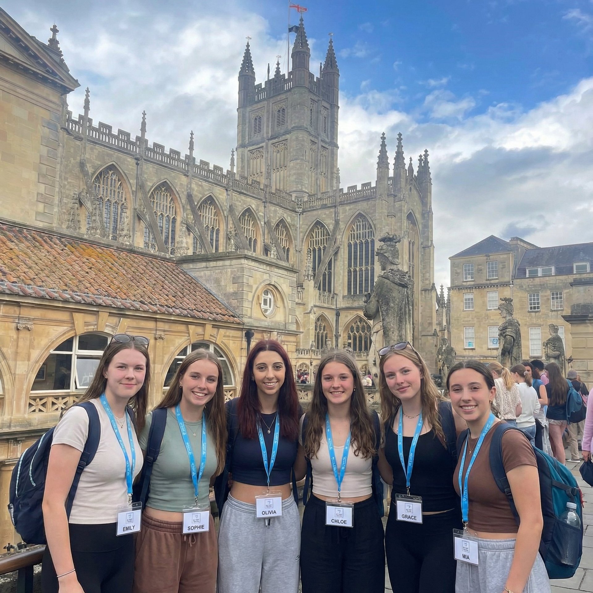 Six young women wearing visitor badges smile at camera in front of a Gothic cathedral with stone architecture