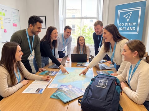 Team of study abroad professionals gathered around a wooden table with maps and materials in a bright office with Go Study Ireland poster
