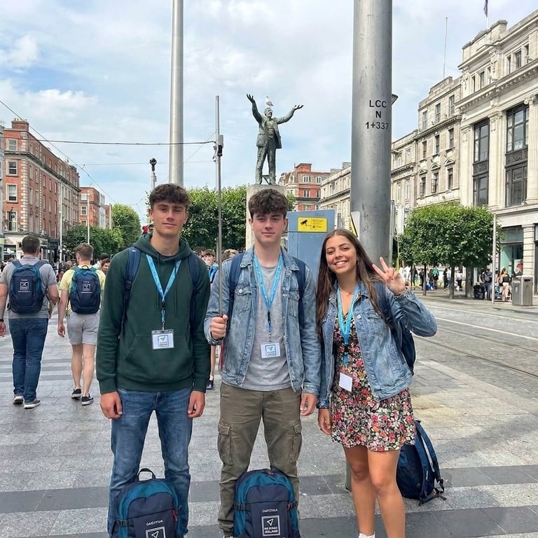 Three young people with backpacks posing in front of a statue in an urban plaza with historic buildings