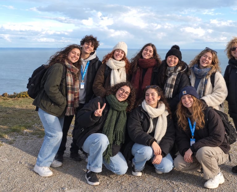 Group of nine young people posing together outdoors by a coastal cliff overlooking the ocean on a cloudy day