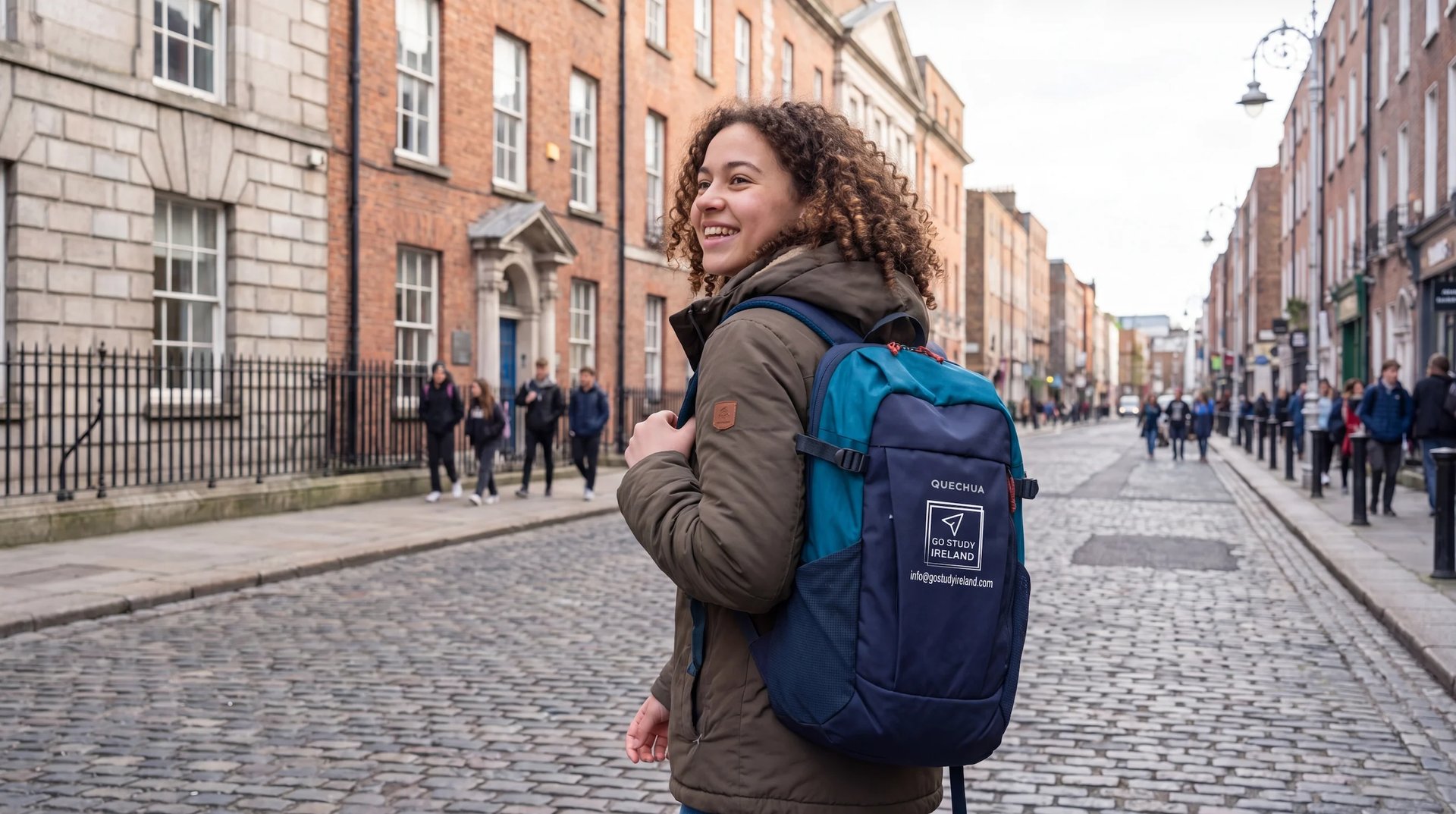 Woman with curly hair wearing a blue and navy backpack smiling on a historic cobblestone street lined with brick buildings